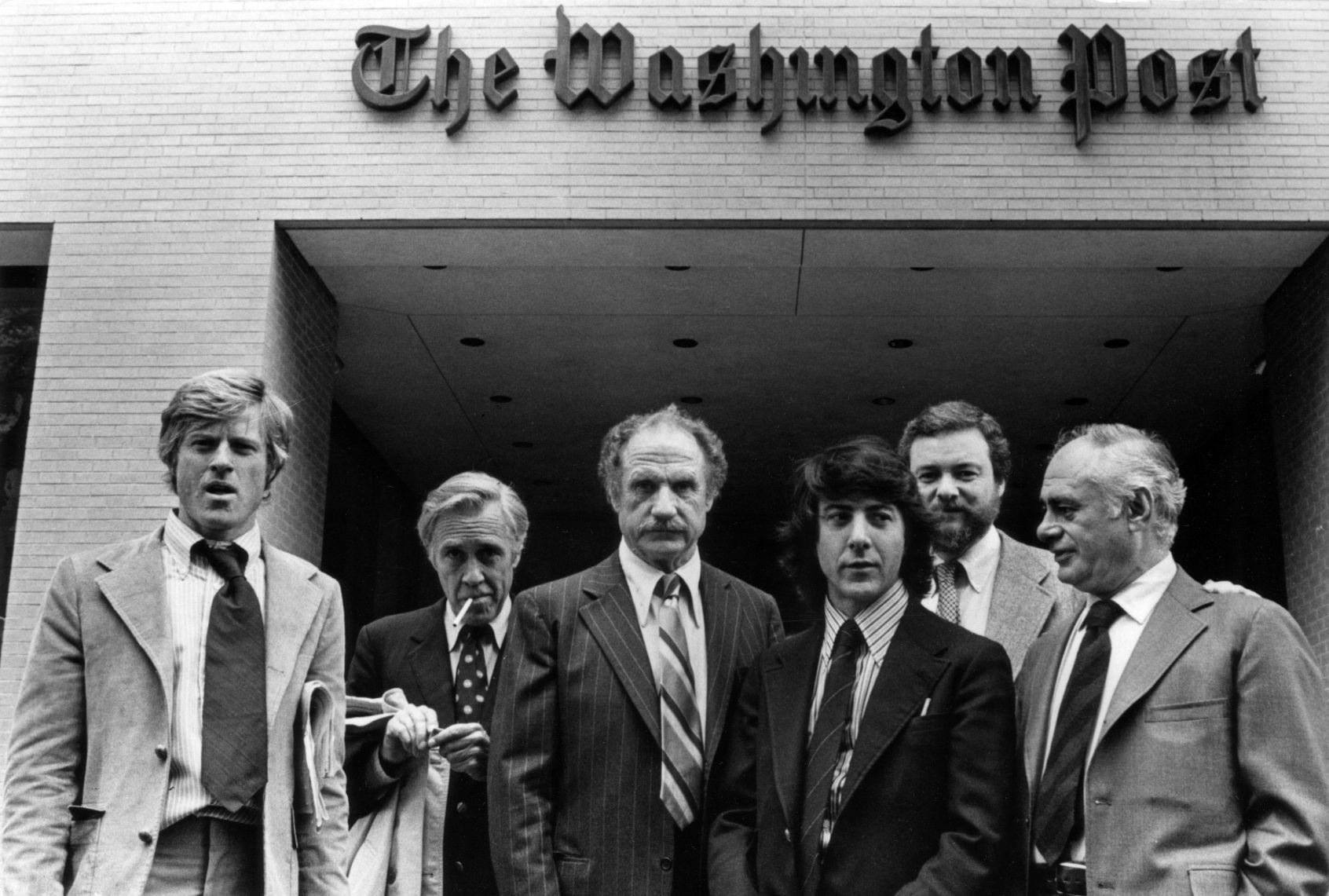 Black and white photo of six men in business suits standing in front of The Washington Post building in the 1970s